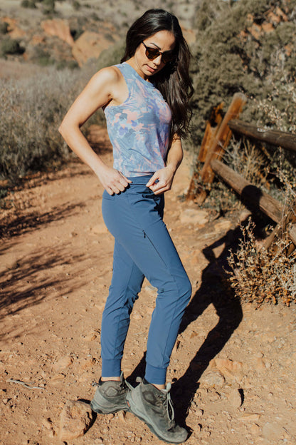 A woman with long, wavy brown hair stands and smiles, wearing a sleeveless lavender High Country Crop Top from Belong Designs and black shorts, paired with white athletic shoes. She has one hand on her hip and is looking to the side against a plain white background. The top is crafted from REPREVE® Certified Recycled Polyester for eco-friendly fashion.