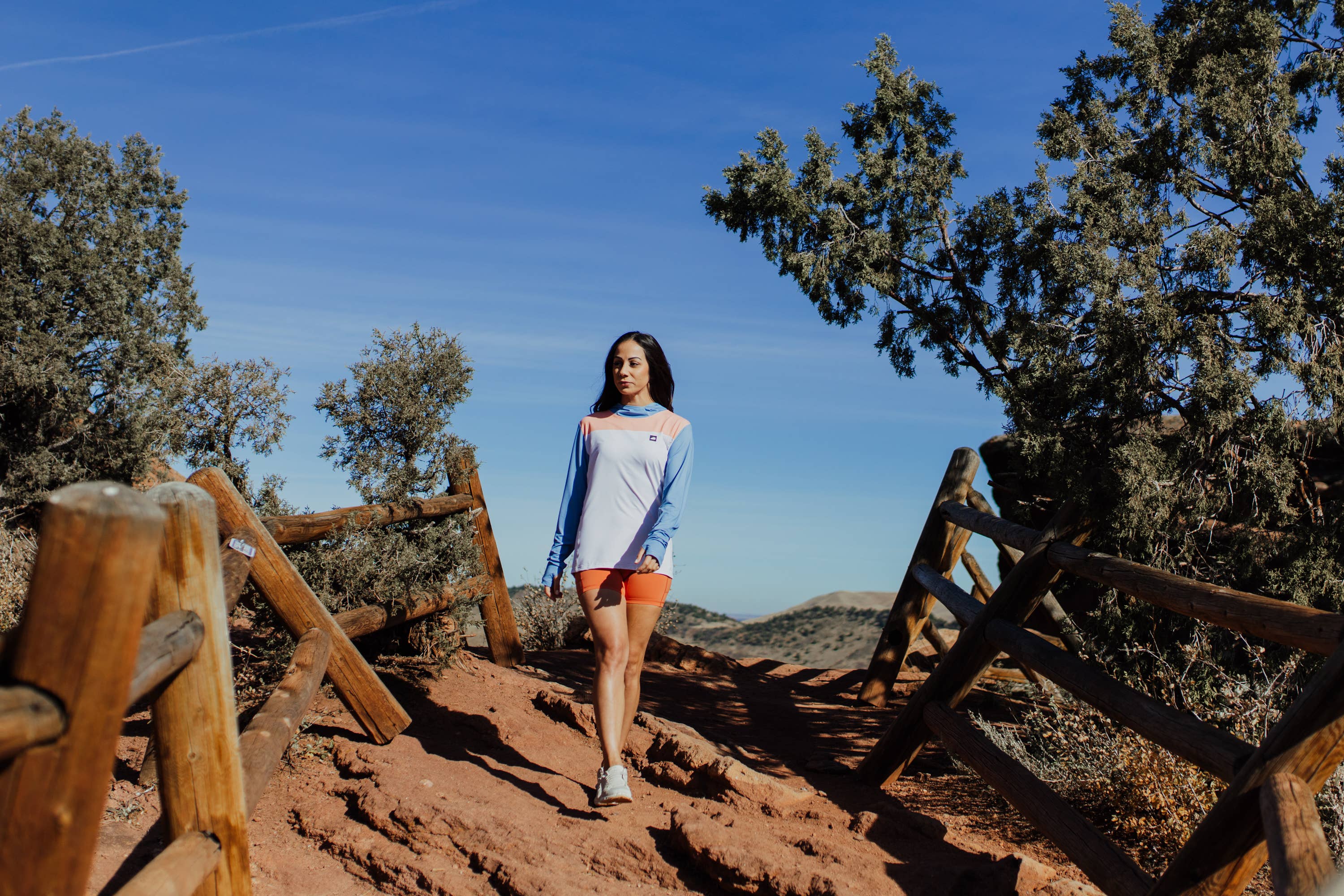 A person in a long-sleeve Sun Hoodie by Belong Designs, featuring a color-blocked design with white, maroon, and gray sections, a black logo on the chest, and wearing dark green shorts stands with hands in pockets. The background is plain white.