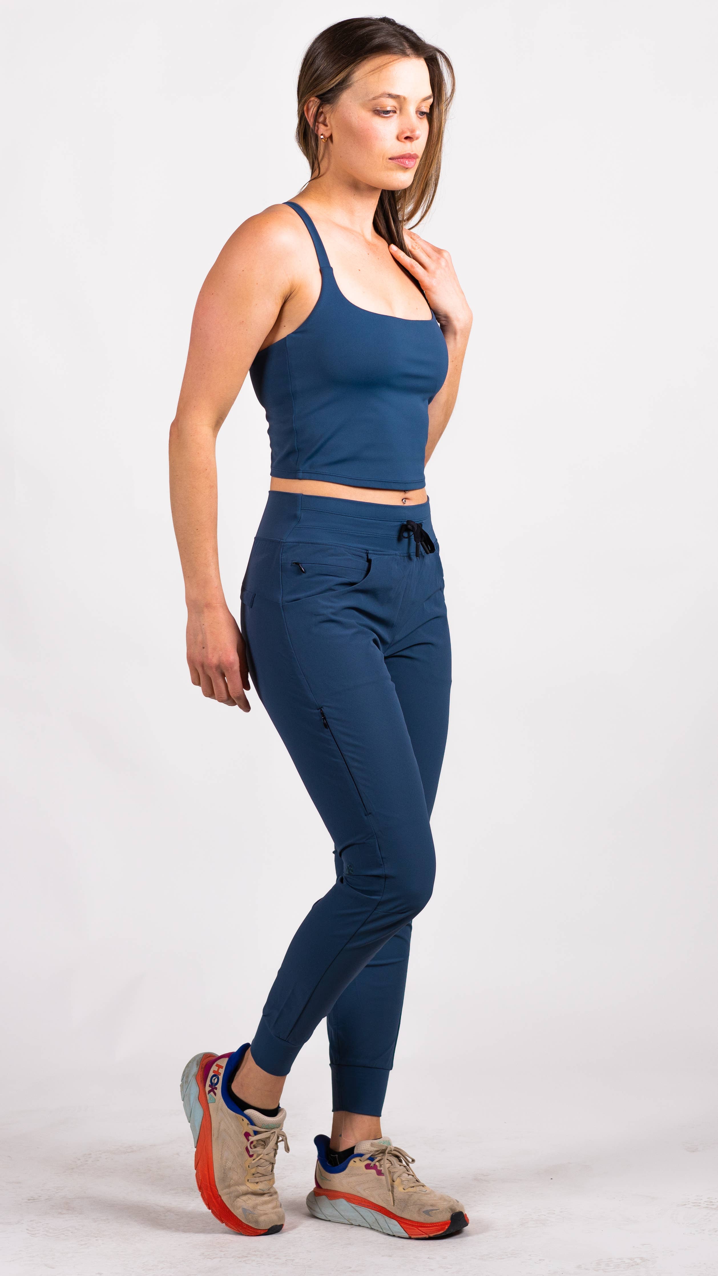 A person with long hair is dressed in a blue workout outfit from Belong Designs, consisting of the Trailhead Tek Tank in Steel Blue with adjustable straps and jogger-style pants. They are posing against a simple, white background, wearing sneakers. The person is looking to the side with one hand lightly resting near their neck.