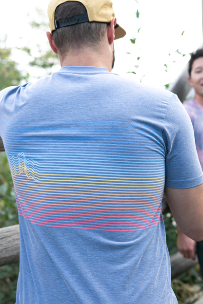 A man stands against a plain background, smiling. He wears a blue and orange camo-patterned Traverse Tek Tee from Belong Designs, made from Recycled VersaTek™ and REPREVE® polyester, black shorts, and brown shoes. He also has a light brown cap. His arms hang relaxed by his sides.