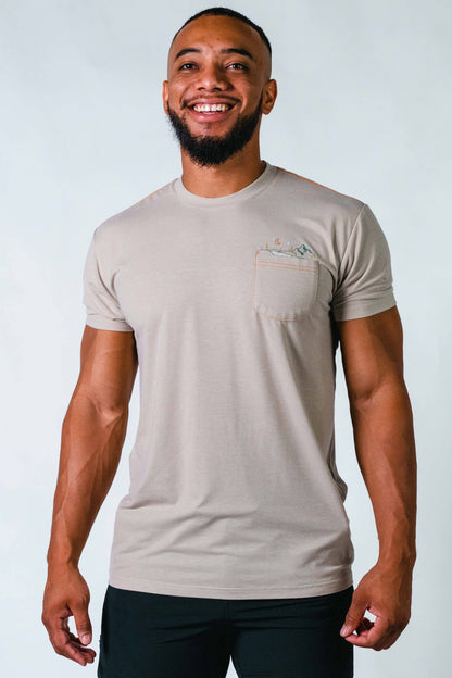 A man stands against a plain background, smiling. He wears a blue and orange camo-patterned Traverse Tek Tee from Belong Designs, made from Recycled VersaTek™ and REPREVE® polyester, black shorts, and brown shoes. He also has a light brown cap. His arms hang relaxed by his sides.