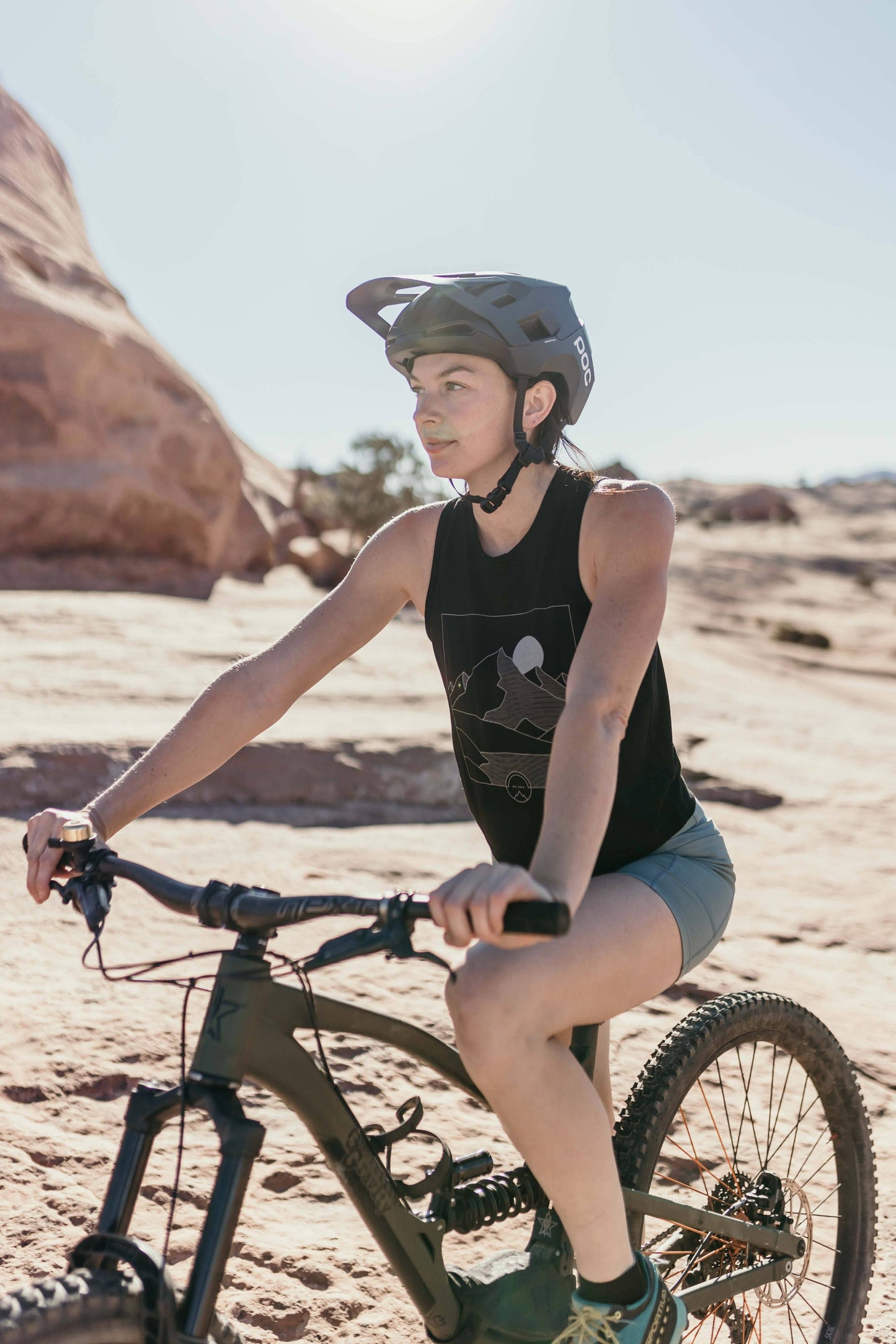 A woman with long, wavy brown hair stands and smiles, wearing a sleeveless lavender High Country Crop Top from Belong Designs and black shorts, paired with white athletic shoes. She has one hand on her hip and is looking to the side against a plain white background. The top is crafted from REPREVE® Certified Recycled Polyester for eco-friendly fashion.