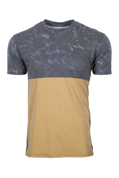 A man stands against a plain background, smiling. He wears a blue and orange camo-patterned Traverse Tek Tee from Belong Designs, made from Recycled VersaTek™ and REPREVE® polyester, black shorts, and brown shoes. He also has a light brown cap. His arms hang relaxed by his sides.