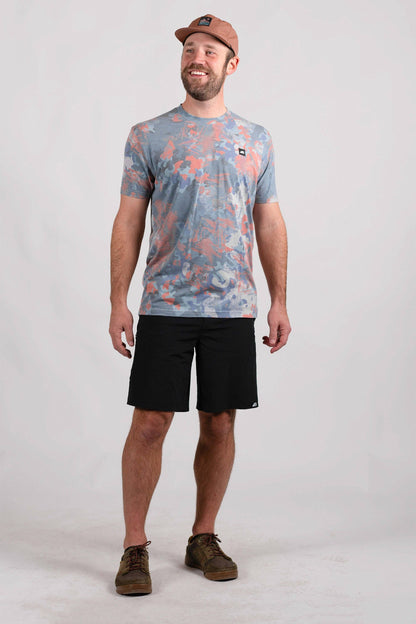 A man stands against a plain background, smiling. He wears a blue and orange camo-patterned Traverse Tek Tee from Belong Designs, made from Recycled VersaTek™ and REPREVE® polyester, black shorts, and brown shoes. He also has a light brown cap. His arms hang relaxed by his sides.