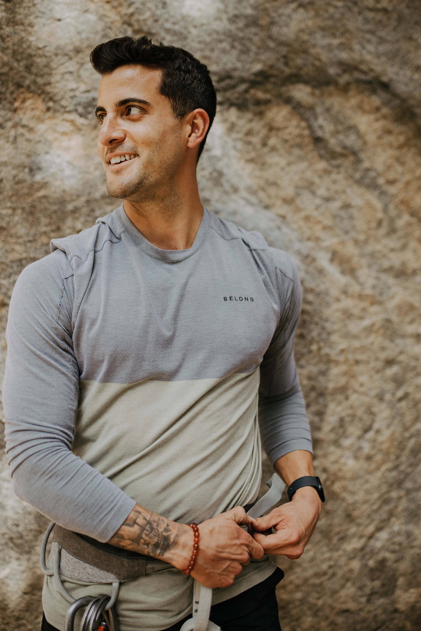 A bearded man with short hair is wearing a Men's Base Camp Tek Long Sleeve shirt from Belong Designs. The shirt, in the Green Block colorway, features "BELONG" printed on the chest and has a two-toned design with a grey top half and an olive green bottom half. Crafted from REPREVE® certified recycled polyester, he stands against a plain, light background.