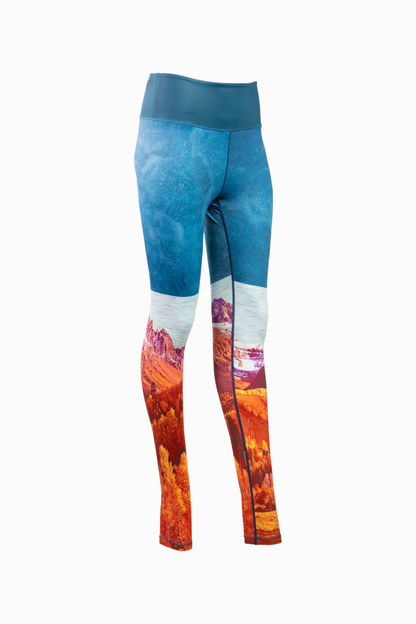 A pair of women's Wayfarer Leggings from Belong Designs featuring a vivid landscape design. The bottom half showcases rocky mountains in red and orange hues, transitioning to blue and white at the top, resembling a sky. Made from breathable PrimaFlex material, the leggings have a fitted style and a solid teal waistband.