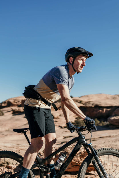 A man stands against a plain background, smiling. He wears a blue and orange camo-patterned Traverse Tek Tee from Belong Designs, made from Recycled VersaTek™ and REPREVE® polyester, black shorts, and brown shoes. He also has a light brown cap. His arms hang relaxed by his sides.