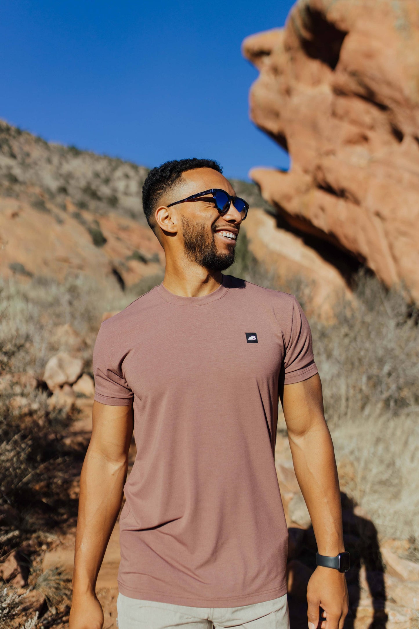 A man stands on a rocky trail with mountainous terrain in the background, wearing the mauve Men's Core Traverse Tek Tee from Belong Designs, light gray athletic fit shorts, dark socks, black shoes, and reflective sunglasses. The sky is clear and blue, with dry shrubs surrounding the trail.