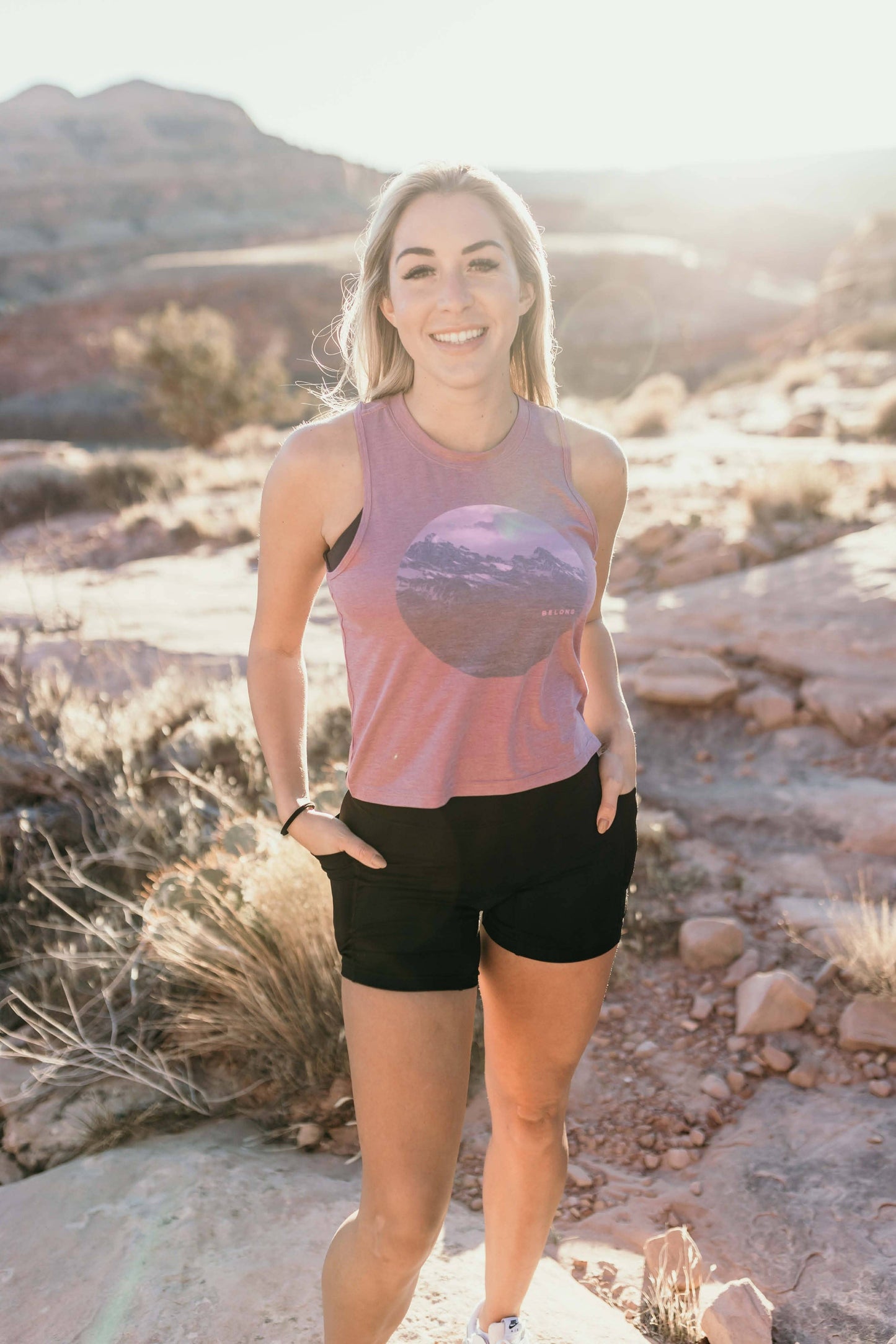 A woman with long, wavy brown hair stands and smiles, wearing a sleeveless lavender High Country Crop Top from Belong Designs and black shorts, paired with white athletic shoes. She has one hand on her hip and is looking to the side against a plain white background. The top is crafted from REPREVE® Certified Recycled Polyester for eco-friendly fashion.