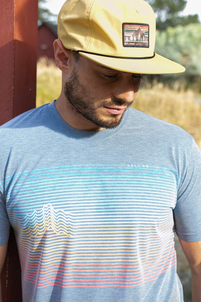 A man stands against a plain background, smiling. He wears a blue and orange camo-patterned Traverse Tek Tee from Belong Designs, made from Recycled VersaTek™ and REPREVE® polyester, black shorts, and brown shoes. He also has a light brown cap. His arms hang relaxed by his sides.