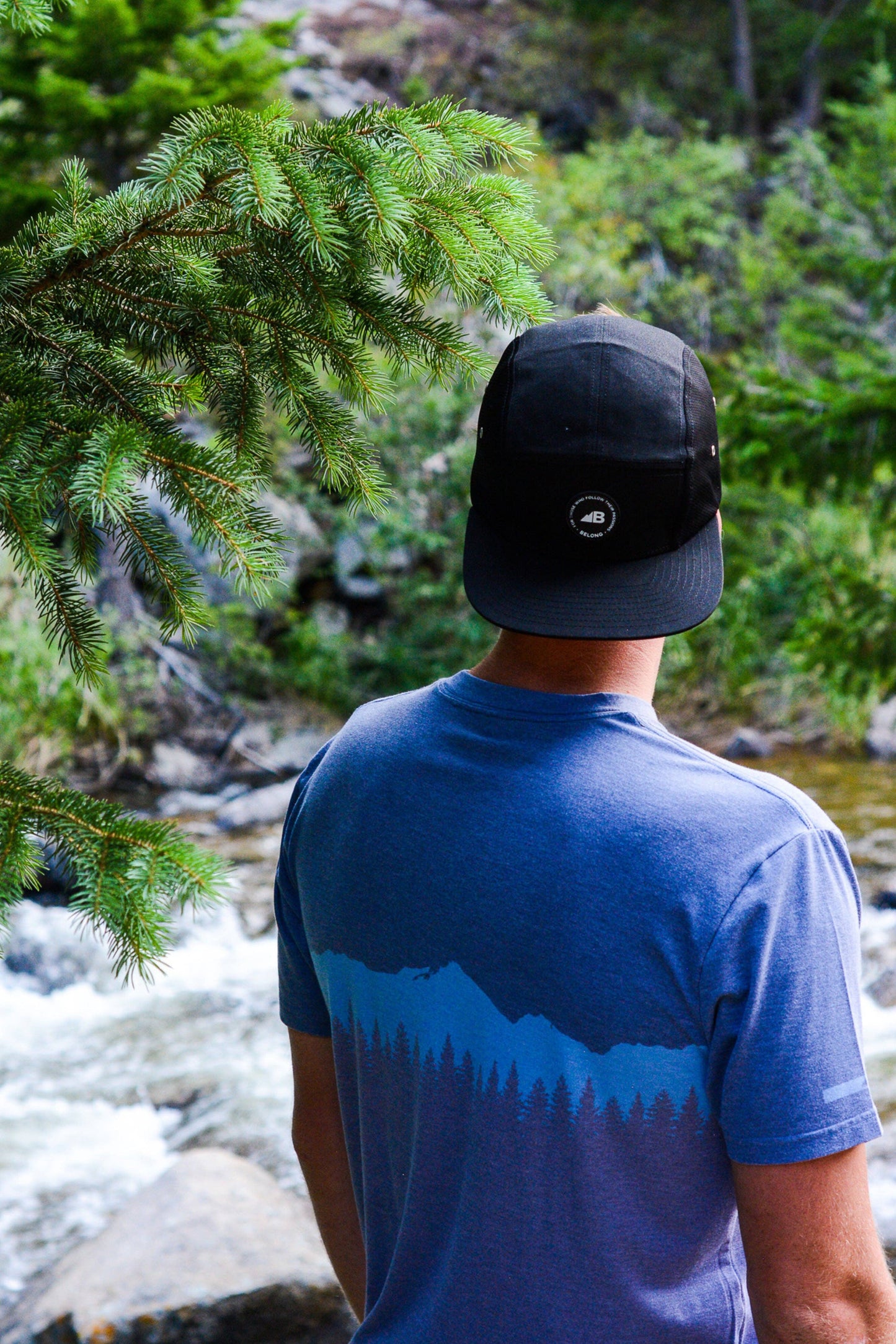 A man stands against a plain background, smiling. He wears a blue and orange camo-patterned Traverse Tek Tee from Belong Designs, made from Recycled VersaTek™ and REPREVE® polyester, black shorts, and brown shoes. He also has a light brown cap. His arms hang relaxed by his sides.