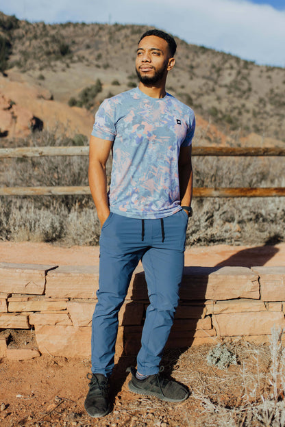 A man stands against a plain background, smiling. He wears a blue and orange camo-patterned Traverse Tek Tee from Belong Designs, made from Recycled VersaTek™ and REPREVE® polyester, black shorts, and brown shoes. He also has a light brown cap. His arms hang relaxed by his sides.