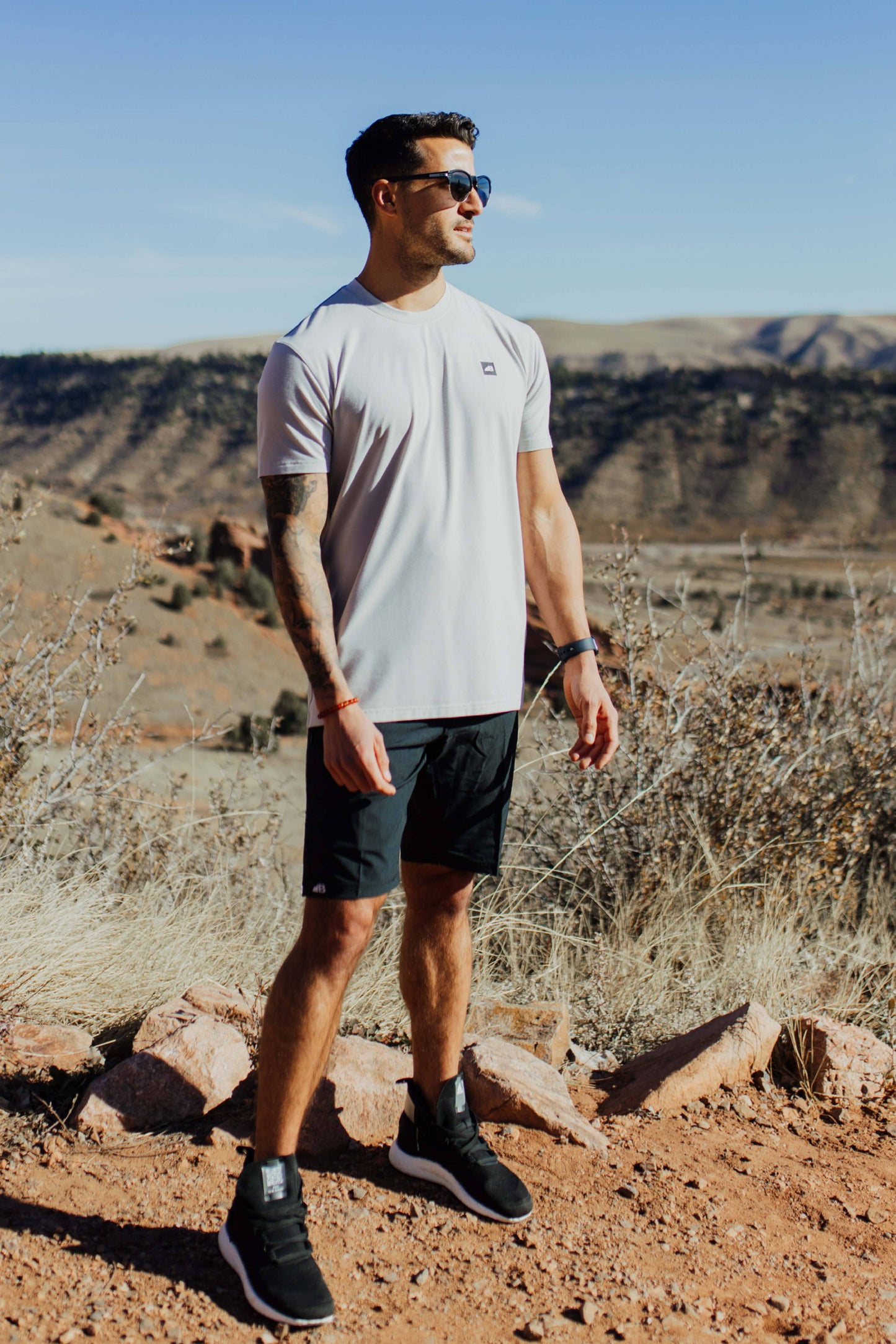 A man stands on a rocky trail with mountainous terrain in the background, wearing the mauve Men's Core Traverse Tek Tee from Belong Designs, light gray athletic fit shorts, dark socks, black shoes, and reflective sunglasses. The sky is clear and blue, with dry shrubs surrounding the trail.