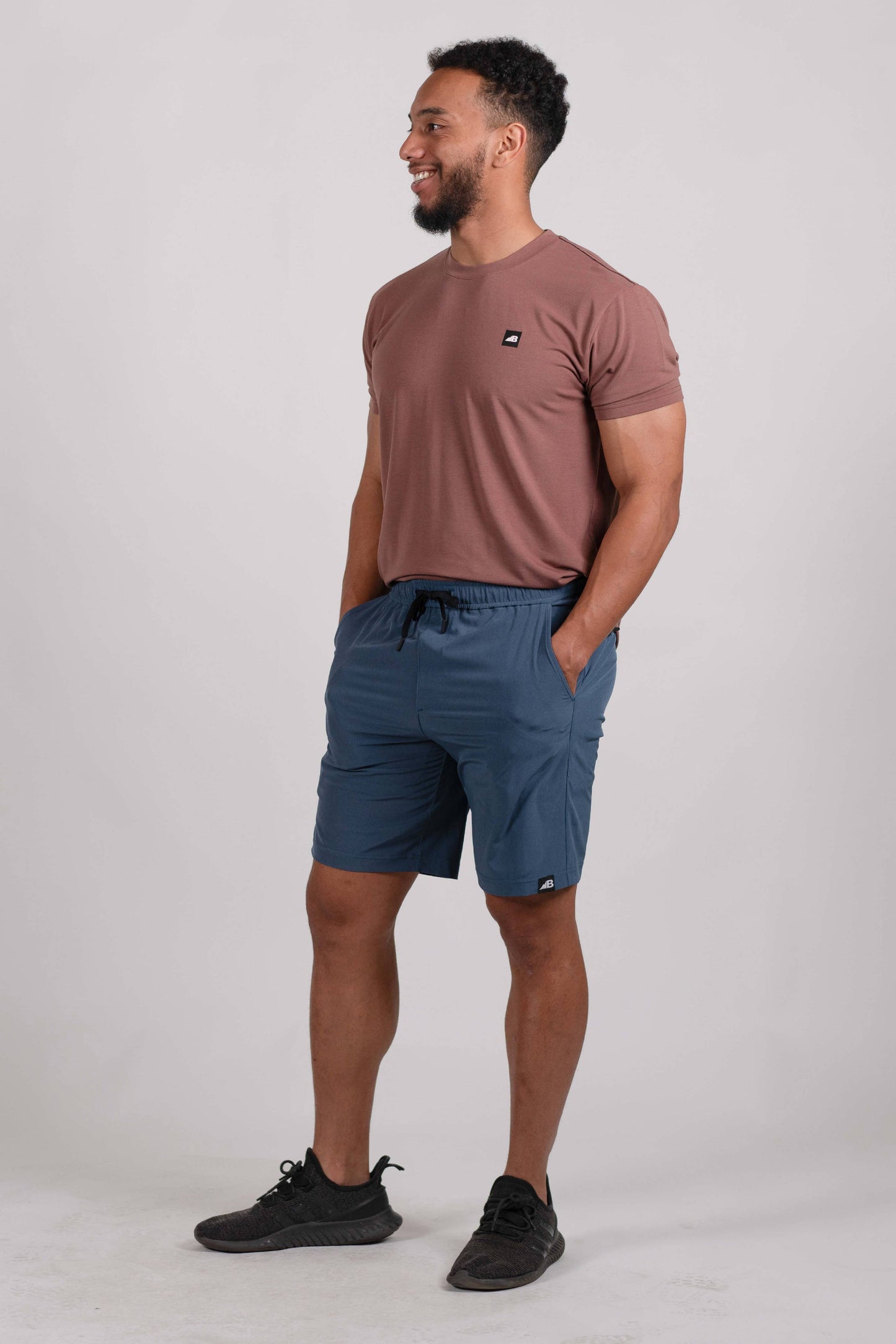 A man stands on a rocky trail with mountainous terrain in the background, wearing the mauve Men's Core Traverse Tek Tee from Belong Designs, light gray athletic fit shorts, dark socks, black shoes, and reflective sunglasses. The sky is clear and blue, with dry shrubs surrounding the trail.