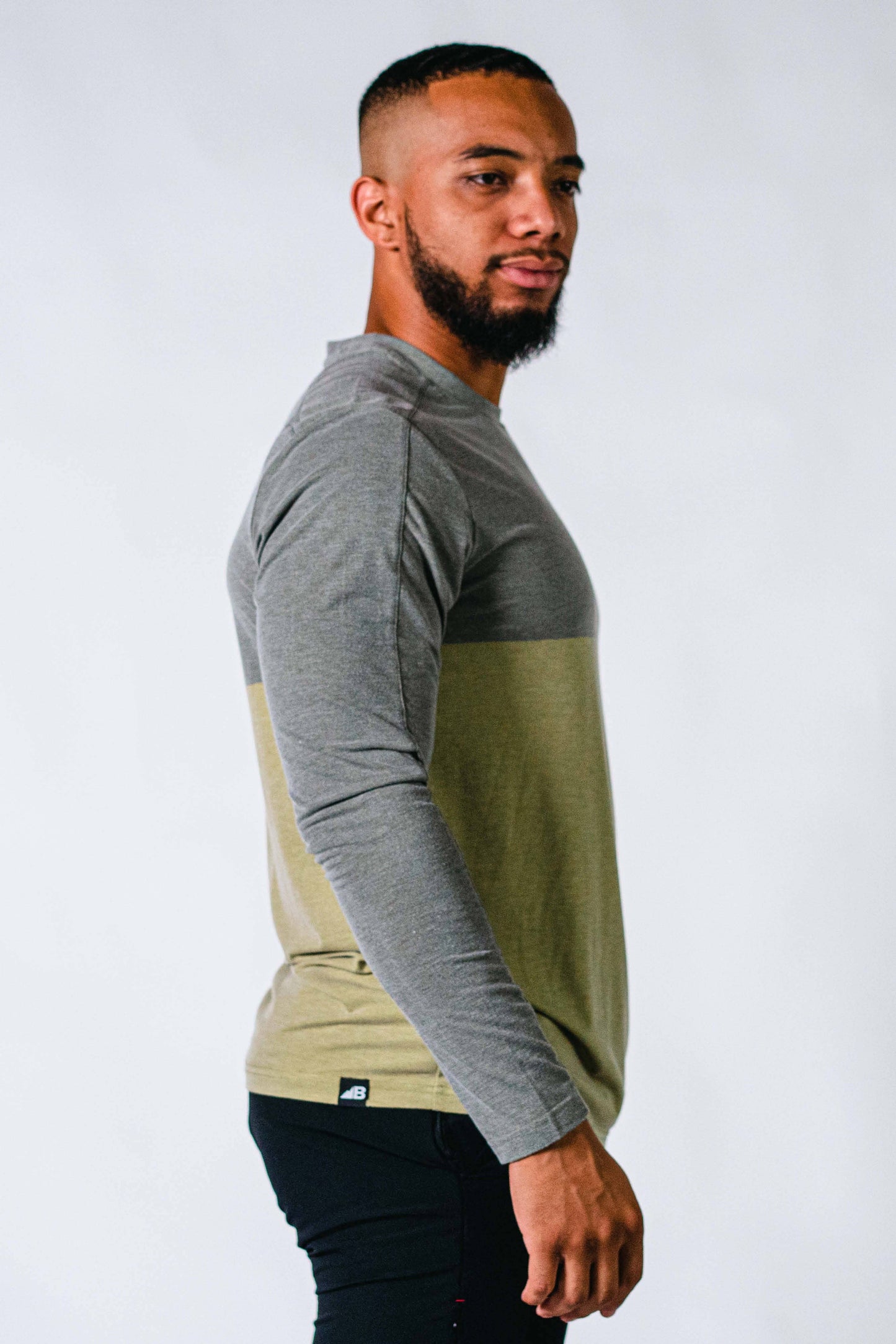 A bearded man with short hair is wearing a Men's Base Camp Tek Long Sleeve shirt from Belong Designs. The shirt, in the Green Block colorway, features "BELONG" printed on the chest and has a two-toned design with a grey top half and an olive green bottom half. Crafted from REPREVE® certified recycled polyester, he stands against a plain, light background.