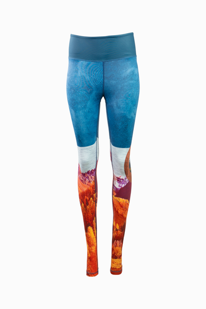 A pair of women's Wayfarer Leggings from Belong Designs featuring a vivid landscape design. The bottom half showcases rocky mountains in red and orange hues, transitioning to blue and white at the top, resembling a sky. Made from breathable PrimaFlex material, the leggings have a fitted style and a solid teal waistband.