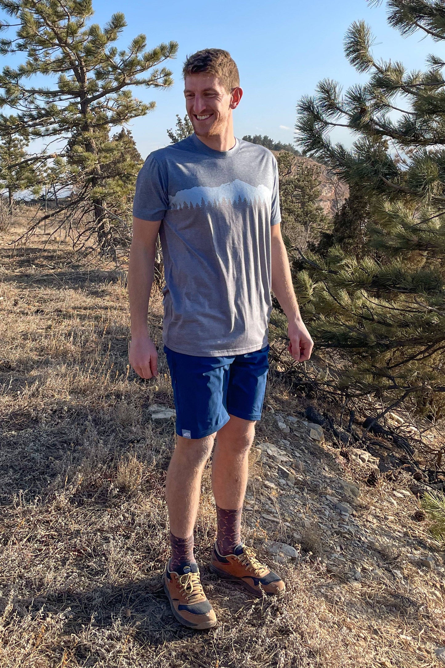 A man stands against a plain background, smiling. He wears a blue and orange camo-patterned Traverse Tek Tee from Belong Designs, made from Recycled VersaTek™ and REPREVE® polyester, black shorts, and brown shoes. He also has a light brown cap. His arms hang relaxed by his sides.