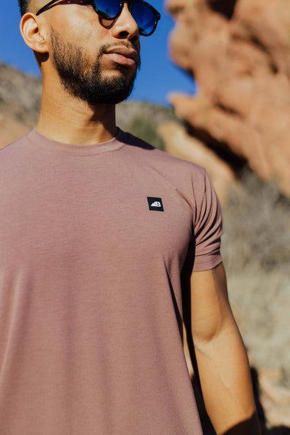 A man stands on a rocky trail with mountainous terrain in the background, wearing the mauve Men's Core Traverse Tek Tee from Belong Designs, light gray athletic fit shorts, dark socks, black shoes, and reflective sunglasses. The sky is clear and blue, with dry shrubs surrounding the trail.