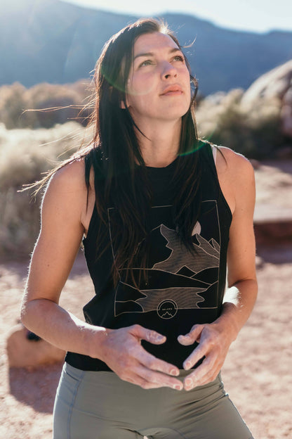 A woman with long, wavy brown hair stands and smiles, wearing a sleeveless lavender High Country Crop Top from Belong Designs and black shorts, paired with white athletic shoes. She has one hand on her hip and is looking to the side against a plain white background. The top is crafted from REPREVE® Certified Recycled Polyester for eco-friendly fashion.