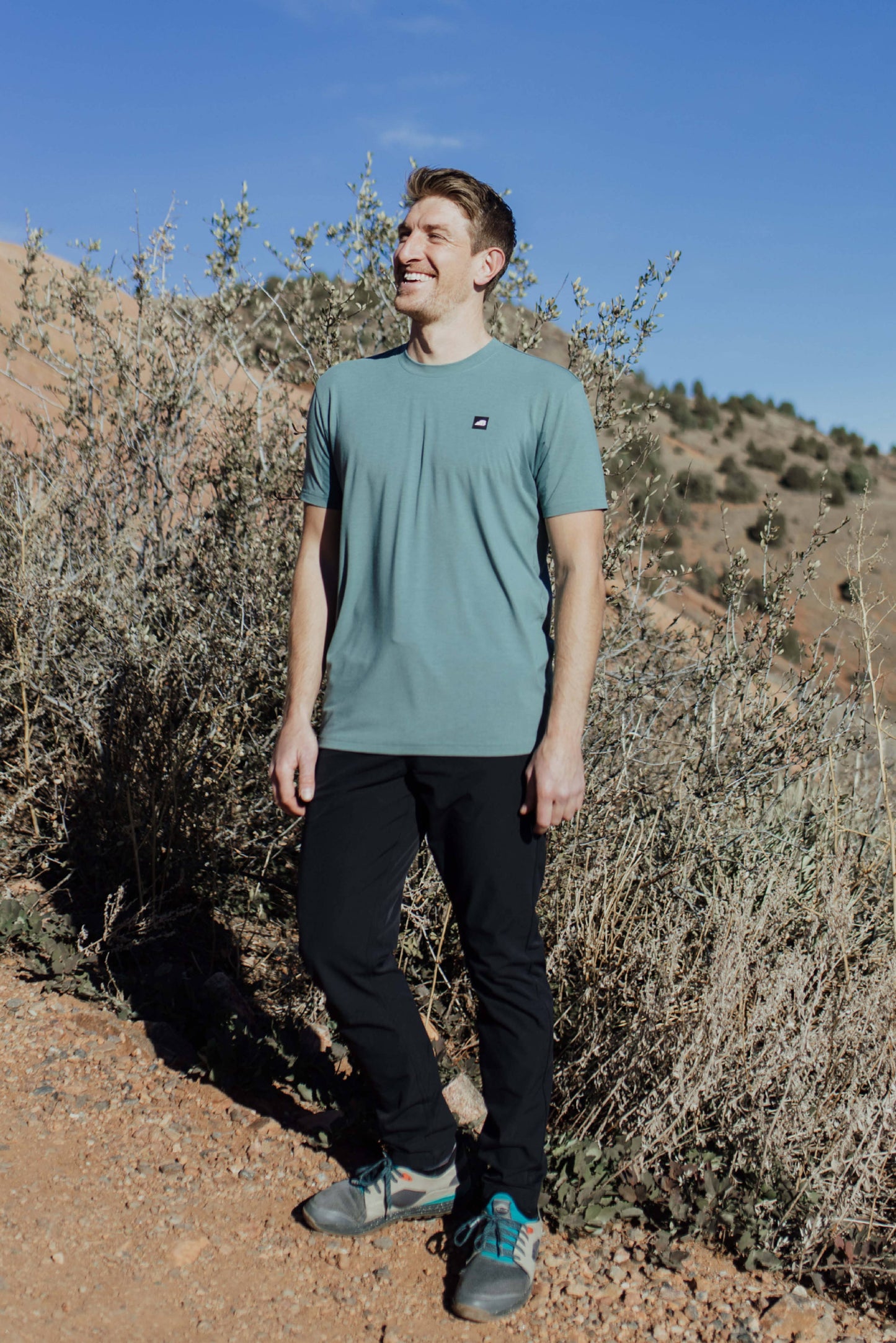 A man stands on a rocky trail with mountainous terrain in the background, wearing the mauve Men's Core Traverse Tek Tee from Belong Designs, light gray athletic fit shorts, dark socks, black shoes, and reflective sunglasses. The sky is clear and blue, with dry shrubs surrounding the trail.