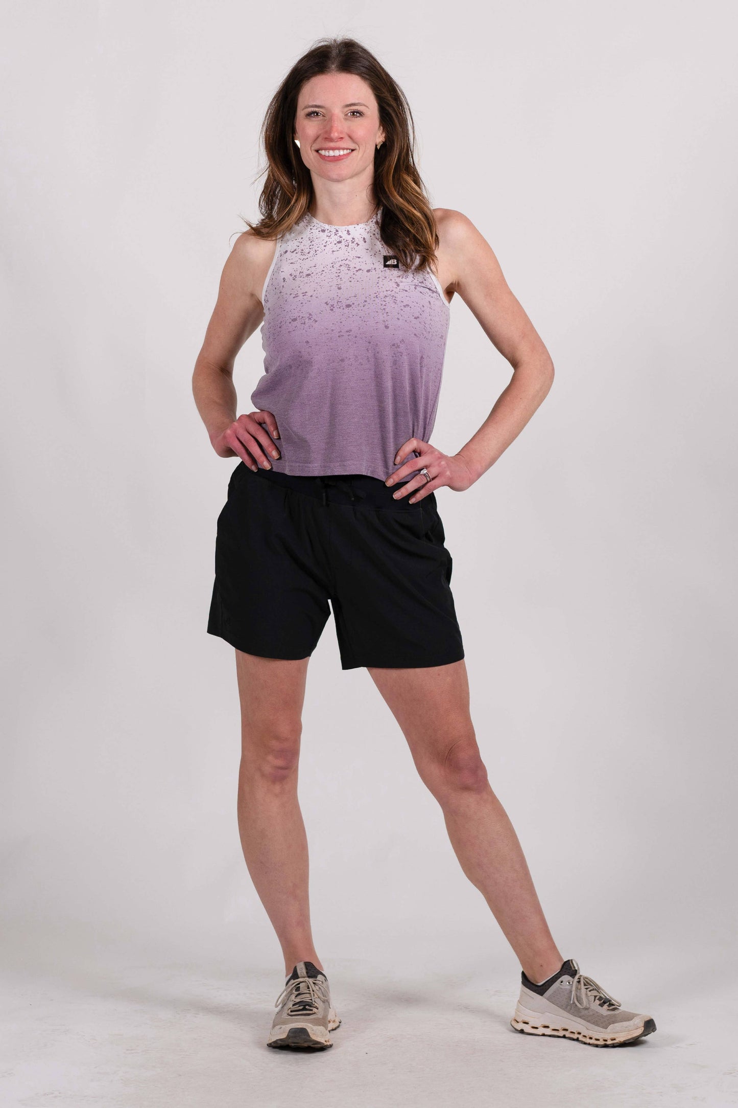 A woman with long, wavy brown hair stands and smiles, wearing a sleeveless lavender High Country Crop Top from Belong Designs and black shorts, paired with white athletic shoes. She has one hand on her hip and is looking to the side against a plain white background. The top is crafted from REPREVE® Certified Recycled Polyester for eco-friendly fashion.