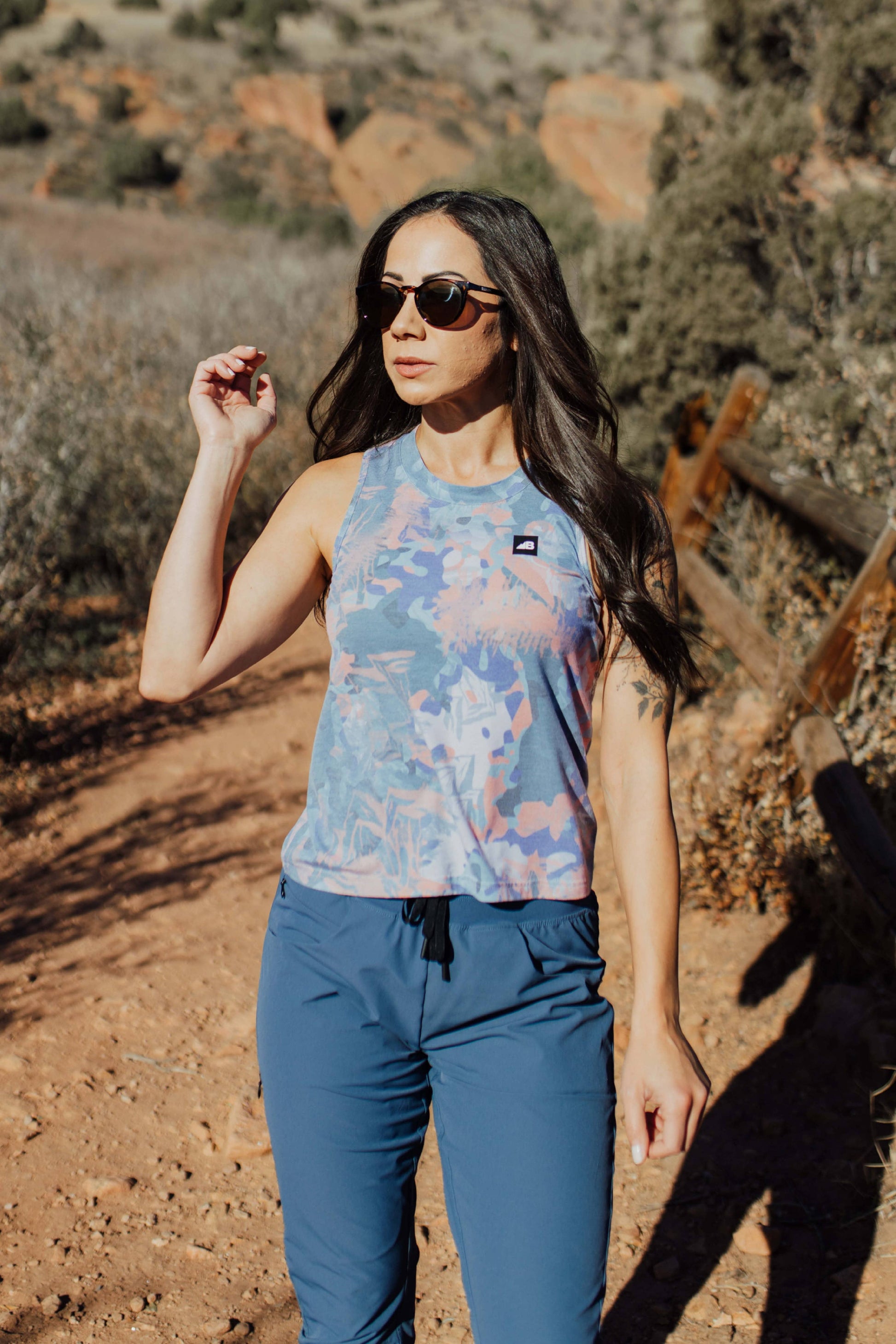 A woman with long, wavy brown hair stands and smiles, wearing a sleeveless lavender High Country Crop Top from Belong Designs and black shorts, paired with white athletic shoes. She has one hand on her hip and is looking to the side against a plain white background. The top is crafted from REPREVE® Certified Recycled Polyester for eco-friendly fashion.