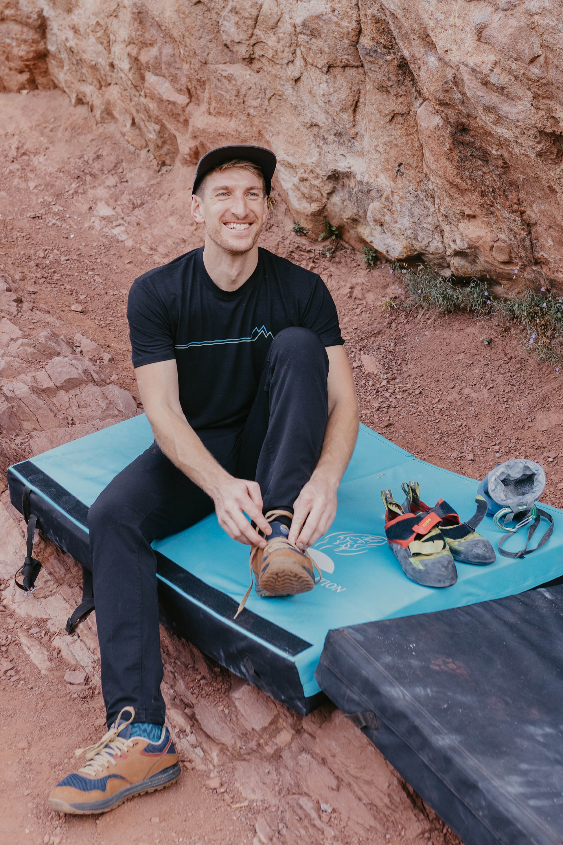 A man stands against a plain background, smiling. He wears a blue and orange camo-patterned Traverse Tek Tee from Belong Designs, made from Recycled VersaTek™ and REPREVE® polyester, black shorts, and brown shoes. He also has a light brown cap. His arms hang relaxed by his sides.