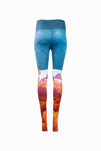 A pair of women's Wayfarer Leggings from Belong Designs featuring a vivid landscape design. The bottom half showcases rocky mountains in red and orange hues, transitioning to blue and white at the top, resembling a sky. Made from breathable PrimaFlex material, the leggings have a fitted style and a solid teal waistband.