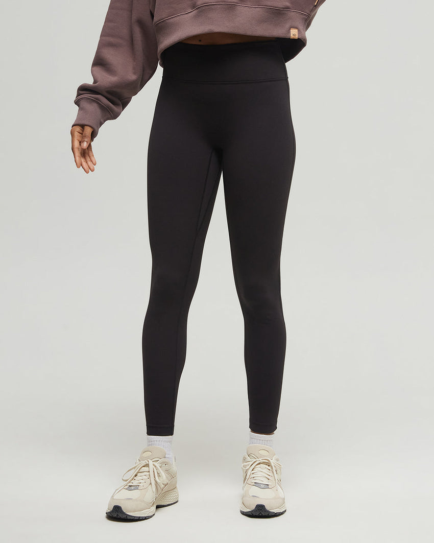 A person wearing a cropped dark sweatshirt made from recycled materials, high-waisted black Tentree inMotion Brushed Leggings, and white sneakers stands against a plain light gray background. Only the torso and legs are visible. The person's hands are slightly outstretched by their sides.