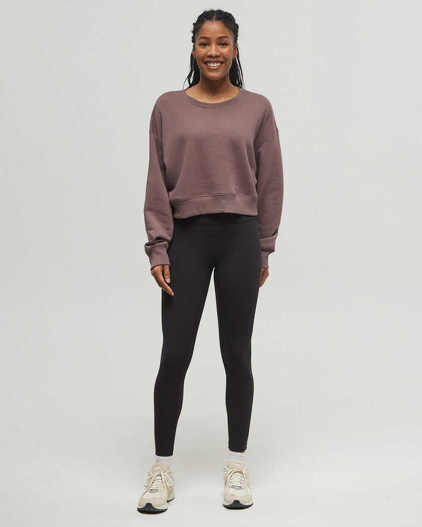 A person wearing a cropped dark sweatshirt made from recycled materials, high-waisted black Tentree inMotion Brushed Leggings, and white sneakers stands against a plain light gray background. Only the torso and legs are visible. The person's hands are slightly outstretched by their sides.