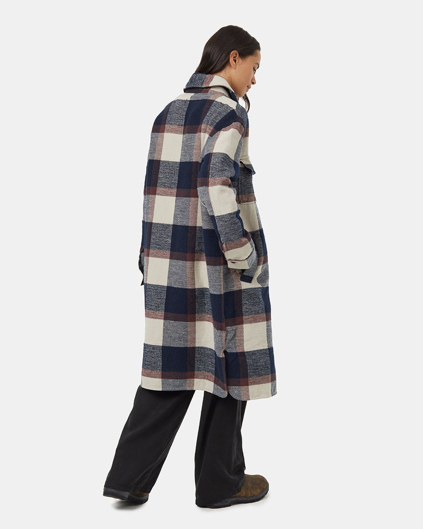 A person wearing the Tentree Heavy Weight Flannel Long Jacket made from recycled materials, featuring large blue, white, grey, and burgundy checks. With one hand in a pocket and the other resting by their side, they stand against a plain background with long hair flowing down.