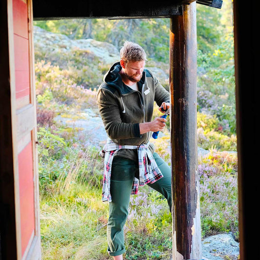 A man stands on a porch set amidst nature, clad in green pants and a dark Men's Skauen Full Zip Hoodie from Amundsen, with a flannel shirt tied around his waist. He seems focused as he inspects or works on a tool or device in his hands. The scene is framed by lush greenery and rocks in the background, enhancing the tranquil atmosphere.