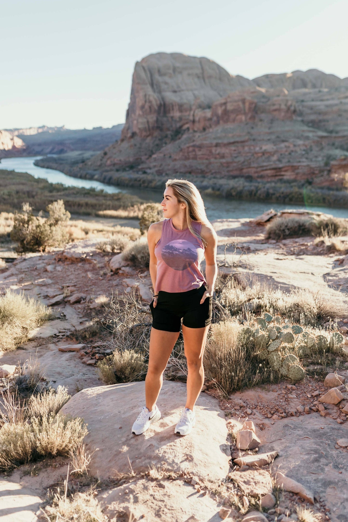 A woman with long, wavy brown hair stands and smiles, wearing a sleeveless lavender High Country Crop Top from Belong Designs and black shorts, paired with white athletic shoes. She has one hand on her hip and is looking to the side against a plain white background. The top is crafted from REPREVE® Certified Recycled Polyester for eco-friendly fashion.