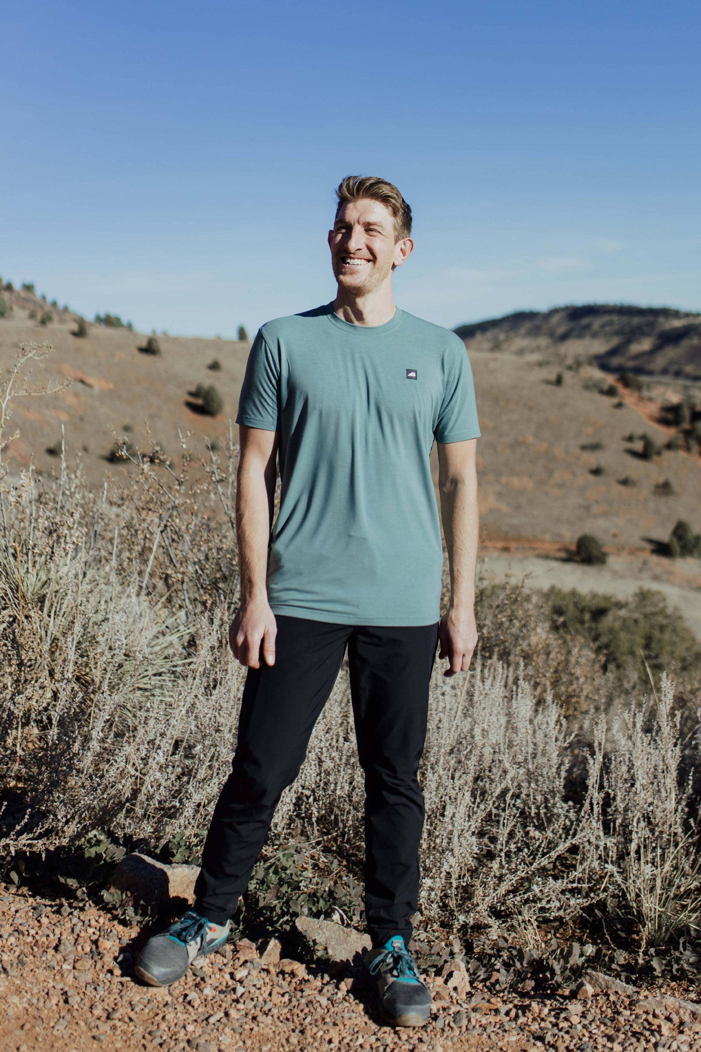 A man stands on a rocky trail with mountainous terrain in the background, wearing the mauve Men's Core Traverse Tek Tee from Belong Designs, light gray athletic fit shorts, dark socks, black shoes, and reflective sunglasses. The sky is clear and blue, with dry shrubs surrounding the trail.