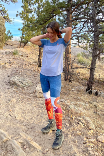 A pair of women's Wayfarer Leggings from Belong Designs featuring a vivid landscape design. The bottom half showcases rocky mountains in red and orange hues, transitioning to blue and white at the top, resembling a sky. Made from breathable PrimaFlex material, the leggings have a fitted style and a solid teal waistband.