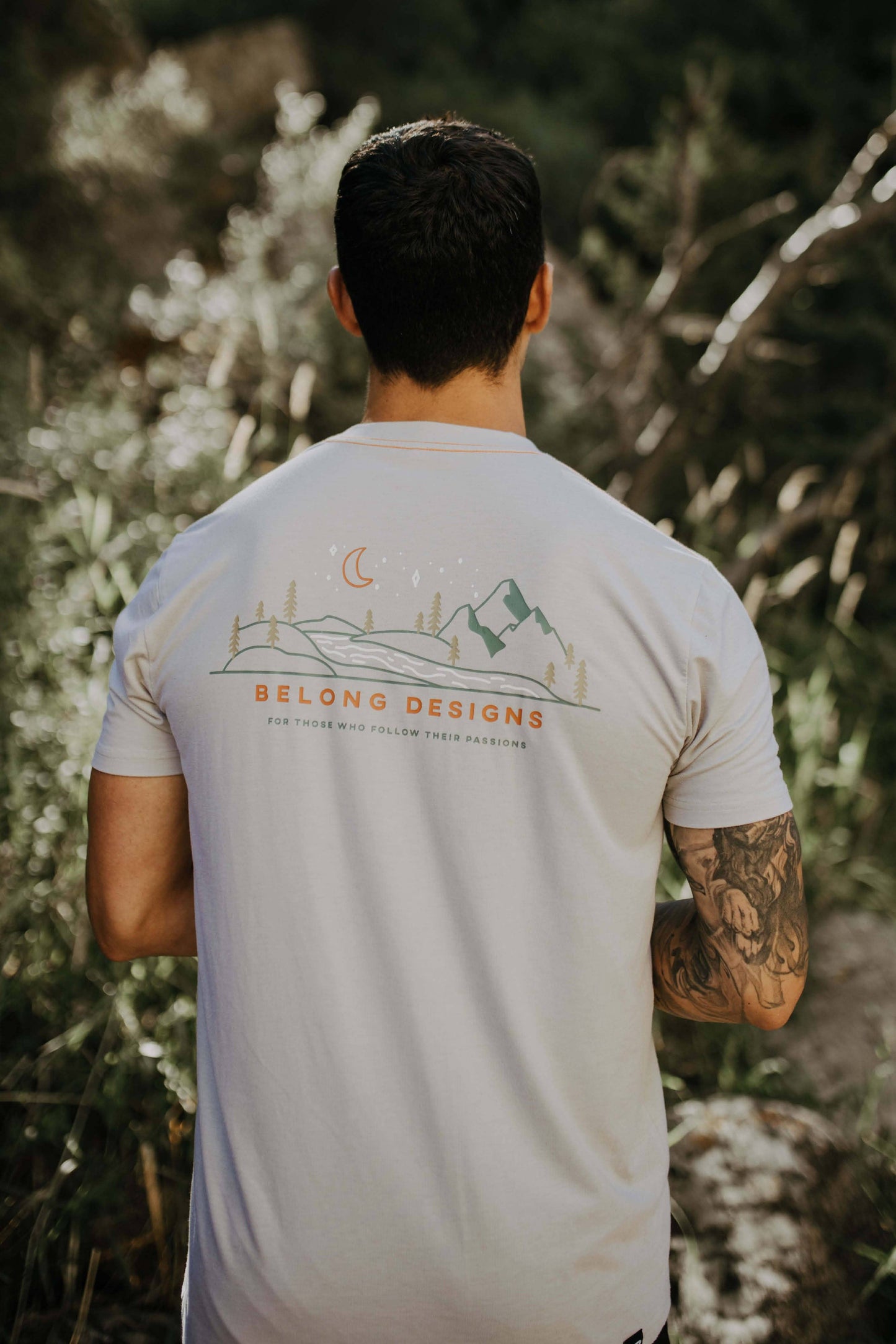 A man stands against a plain background, smiling. He wears a blue and orange camo-patterned Traverse Tek Tee from Belong Designs, made from Recycled VersaTek™ and REPREVE® polyester, black shorts, and brown shoes. He also has a light brown cap. His arms hang relaxed by his sides.