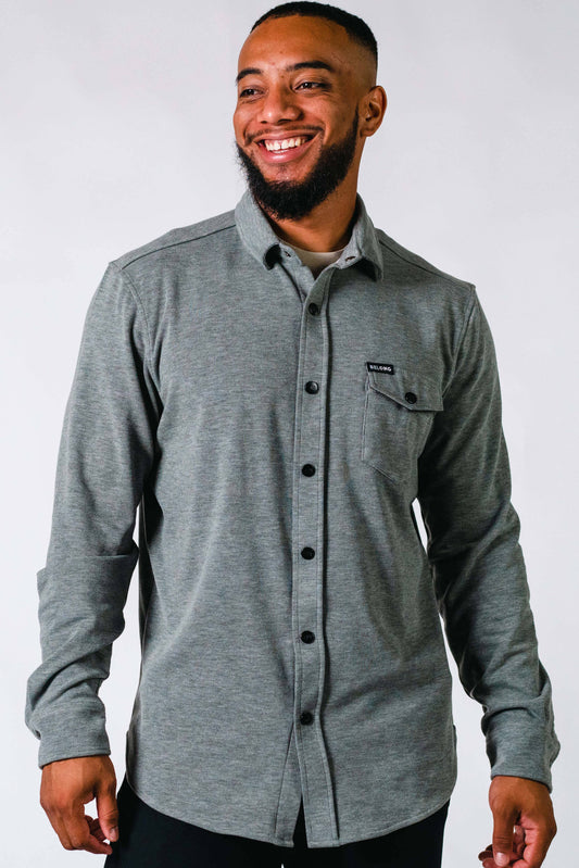 A man with a short beard and a bright smile is standing against a plain background. He is wearing a Sherman Fleece Button Up, long-sleeve, grey shirt by Belong Designs, featuring a pocket on the left side of his chest adorned with a small tag - perfect for a cozy winter weekend.